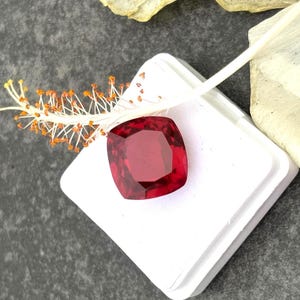Könnte beinhalten: Ein leuchtend roter Edelstein mit Kissenform, präsentiert in einer weißen quadratischen Schachtel. Der Edelstein hat eine tiefrote Farbe und einen hohen Glanz.