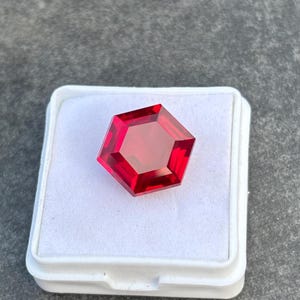 May include: A faceted, pentagon-shaped, deep red gemstone sits in a white plastic case. The gem has a polished, reflective surface and a geometric design. The case is square with a slightly raised lid, against a grey background.