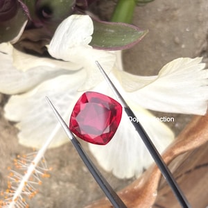 Può includere: Una gemma rosso vibrante con taglio a cuscino, tenuta da una pinzetta in metallo nero. La gemma presenta una superficie sfaccettata che riflette la luce, mostrando una profonda tonalità cremisi. La pietra è posizionata davanti a petali bianchi.