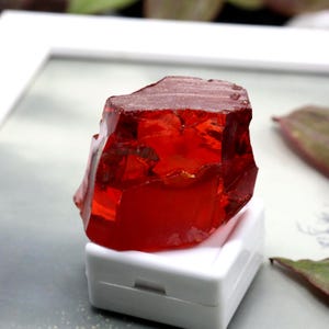 Op de afbeelding: Een levendige, ruwe, rode edelsteen met een kristalstructuur. De edelsteen wordt gepresenteerd op een kleine, witte, rechthoekige doos. De kleur van de edelsteen is een diep, doorschijnend rood, met zichtbare interne facetten.