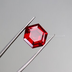 Puede incluir: Una gema facetada, hexagonal, de color rojo intenso, sostenida por unas pinzas plateadas sobre un fondo gris. La gema tiene una superficie pulida y reflectante. El texto "DopeCollectionIN" es visible a la derecha.