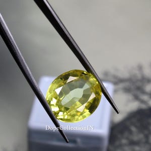May include: A faceted, oval-shaped gemstone in shades of yellow and green is held by black tweezers. The gem is set against a blurred background. The text "DopeCollectionIN" is visible at the bottom.