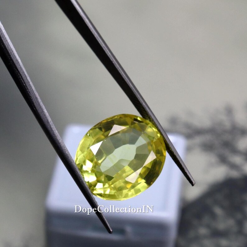 May include: A faceted, oval-shaped gemstone in shades of yellow and green is held by black tweezers. The gem is set against a blurred background. The text "DopeCollectionIN" is visible at the bottom.