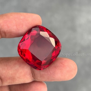 以下が含まれることがあります： 鮮やかな赤色のクッションカットの宝石のクローズアップ。指で宝石を持ち、研磨された表面と内部の反射を強調しています。宝石は角が丸い正方形で、ルビーに似ています。