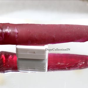 Könnte beinhalten: Ein tiefroter, länglicher, zylindrischer Edelstein mit einer glatten, polierten Oberfläche und einem leicht zulaufenden Ende. Der Stein liegt horizontal auf einem kleinen, rechteckigen weißen Ständer, wobei der Text DopeCollectionIN in der Nähe der Basis sichtbar ist.