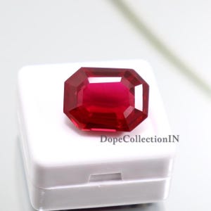 Puede incluir: Una gema de color rojo intenso y vibrante con forma de corte esmeralda, que presenta múltiples facetas que reflejan la luz. La gema está colocada sobre una pequeña caja de presentación cuadrada de plástico blanco con el texto DopeCollectionIN impreso.