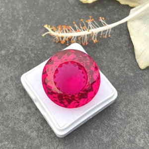 May include: A large, round, faceted, hot pink gemstone is displayed on a white box. The gemstone has a brilliant cut, reflecting light. A white flower with a yellow center is in the background.