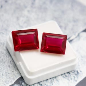 May include: Two rectangular, faceted, deep red gemstones. The gems are set on a white surface, with a light blue and white patterned background. The gems have a vibrant, polished appearance, showcasing their clarity and color.
