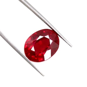 Può includere: Una gemma di rubino sfaccettata, di forma ovale, tenuta da pinzette d'argento. Il rubino è di un colore rosso intenso e vibrante, con molteplici sfaccettature che riflettono la luce. Lo sfondo è bianco.