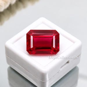 Pode incluir: Um rubi de corte retangular, apresentado numa caixa branca. O rubi é de um vermelho profundo e saturado, com uma superfície polida. A gema é apresentada sobre um fundo branco, realçando o seu brilho.