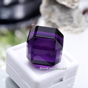 Puede incluir: Una gema facetada de color púrpura intenso con forma geométrica. La gema está colocada sobre una caja de exhibición blanca. El texto "DopeCollectionIN" es visible en la caja. Las facetas de la gema reflejan la luz.