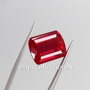 Puede incluir: Una piedra preciosa roja vibrante de corte rectangular facetado, sostenida por pinzas de metal plateado. La piedra presenta una superficie clara y pulida con bordes geométricos definidos y reflejos de luz internos. El texto DopeCollectionIN es visible.