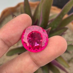 May include: A faceted, round, hot pink gemstone. The gem is held between fingers, with a blurred background of green and purple plant leaves. The gem's cut creates a sparkling effect.