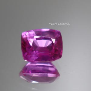 Puede incluir: Una gema rosa, tallada en cojín, con múltiples facetas. La gema se presenta sobre una superficie reflectante, creando un efecto espejo. El color es un rosa vibrante y saturado, con sutiles reflejos en las facetas.