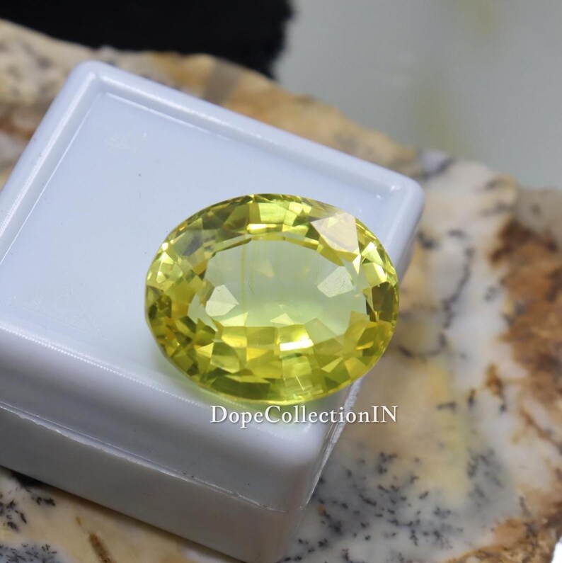 May include: A faceted, oval-shaped, yellow gemstone. The gem is set on a white surface, with the text "DopeCollectionIN" visible. The gemstone's color is a vibrant, light yellow, and it appears to be well-polished.