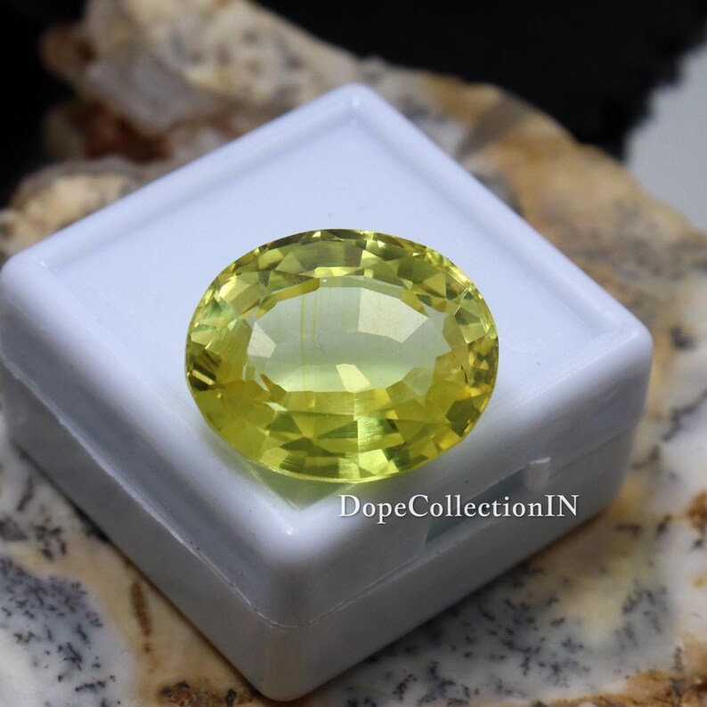 May include: A faceted, oval-shaped, yellow gemstone rests on a white surface. The gem has a brilliant cut, reflecting light. The text "DopeCollectionIN" is visible on the white surface. The gemstone is a vibrant, light yellow color.