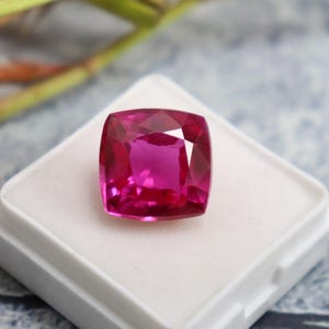 Op de afbeelding: Een levendige roze edelsteen met een vierkante kussen-slijpvorm, getoond op een witte achtergrond.
