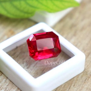 Könnte beinhalten: Ein leuchtend roter Edelstein mit rechteckigem Smaragdschliff, präsentiert in einer weißen quadratischen Box. Der Edelstein ist ein Rubin.