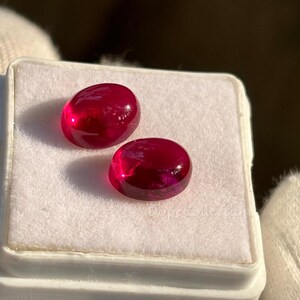 May include: Two oval-shaped, deep red gemstones. The gems are displayed on a white surface inside a clear plastic container. The gems have a polished, reflective surface. The overall theme is jewelry and gemstones.