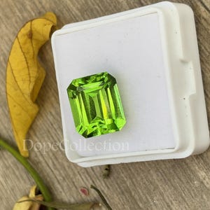 Puede incluir: Una gema verde vibrante con un corte cuadrado de esmeralda, exhibida en una caja blanca. La gema es un peridoto, una variedad de olivino.