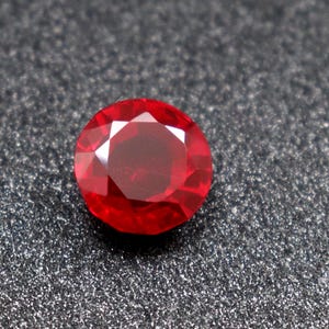 Puede incluir: Una piedra preciosa de rubí rojo intenso con un corte redondo y una superficie facetada.