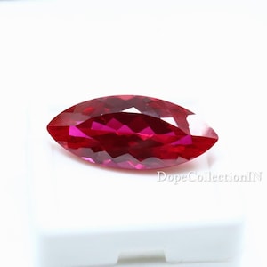 Puede incluir: Una piedra preciosa de corte marquesa con un color rojo intenso y vibrante y una superficie facetada que refleja la luz. La piedra está colocada sobre una base de exhibición blanca con el texto DopeCollectionIN impreso en la esquina inferior derecha.