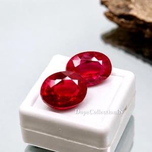 Peut inclure: Deux pierres précieuses rouges de forme ovale sont présentées sur une surface blanche. Les gemmes ont un éclat élevé et sont posées sur un fond neutre. Les gemmes sont probablement des rubis. L'image est bien éclairée.