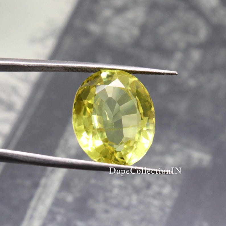 May include: A faceted, oval-shaped, yellow gemstone held by tweezers. The gem has a bright, translucent appearance with multiple internal facets. The background is a blurred, neutral tone. The text "DopeCollectionIN" is visible.