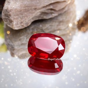 May include: A faceted, oval-shaped, deep red gemstone with a brilliant shine. The gem is reflected on a mirrored surface, with a blurred background of a rock and sparkling lights. The text "DopeCollectionIN" is visible.