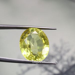 May include: A faceted, oval-shaped, pale yellow gemstone is held by a pair of tweezers. The gem has a brilliant cut, reflecting light. The background is a soft, blurred gray. The text "DopeCollectionIN" is visible at the bottom.