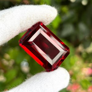 May include: A deep red, rectangular gemstone with a faceted cut, held between two white fabric-covered fingers. The stone displays a rich, translucent crimson hue with sharp, geometric edges and a polished surface that reflects light clearly.