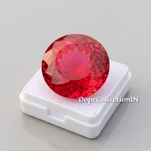 以下が含まれることがあります： 鮮やかな赤色の丸いファセットカットが施された宝石で、ブリリアントカットにより表面に複雑な幾何学的な光の反射が見られます。この宝石は、DopeCollectionINという文字が記された小さな正方形の白いプラスチック製ディスプレイボックスの上に置かれています。