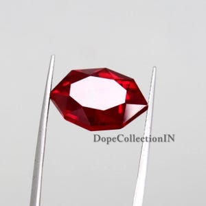 Könnte beinhalten: Ein leuchtend roter, facettierter Edelstein in sechseckiger Form, der von einer silberfarbenen Metallpinzette gehalten wird. Der Edelstein hat eine polierte Oberfläche mit mehreren geometrischen Schliffen. Der Text DopeCollectionIN ist sichtbar.