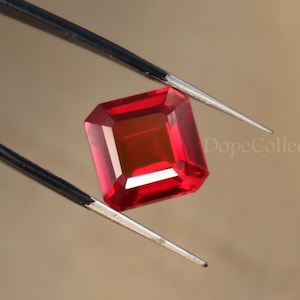 May include: A vibrant red gemstone with a square cut, held by tweezers. The gemstone is clear and reflects light.