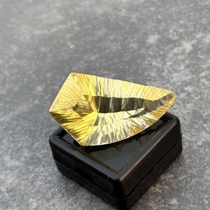 Könnte beinhalten: Ein facettierter, goldgelber Edelstein mit komplizierten Oberflächenmustern, der auf einer schwarzen Vitrine ruht. Der Schliff des Edelsteins erzeugt ein Spiel von Licht und Schatten, das seine Brillanz verstärkt. Die Form ist ein einzigartiges, asymmetrisches Design.