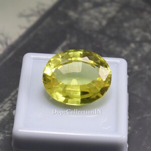 May include: A faceted, oval-shaped, yellow gemstone. The gem is set on a white surface. The text "DopeCollectionIN" is visible on the white surface. The gemstone has a bright, translucent appearance.