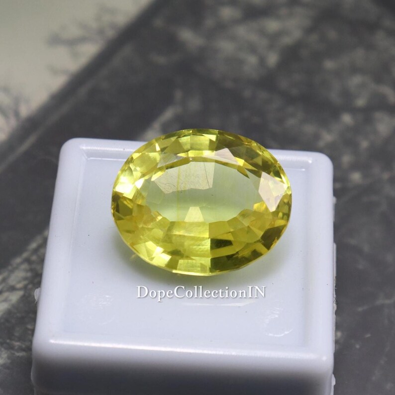 May include: A faceted, oval-shaped, yellow gemstone. The gem is set on a white surface. The text "DopeCollectionIN" is visible on the white surface. The gemstone has a bright, translucent appearance.