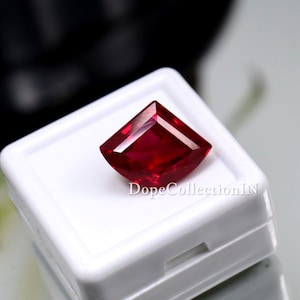 Könnte beinhalten: Ein lebendiger, facettierter roter Edelstein in Drachenform, der in einer weißen Präsentationsbox liegt. Die tiefe Farbe und die scharfen Winkel des Edelsteins werden vor dem neutralen Hintergrund hervorgehoben. Auf der Box steht "DopeCollectionIN".