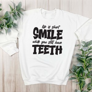 May include: White sweatshirt with black text that reads "Life is short, Smile while you still have Teeth".
