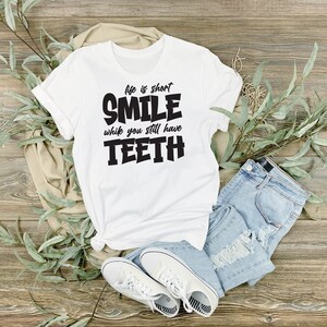 May include: White t-shirt with black text that reads "Life is short, Smile while you still have Teeth". The shirt is laid out on a wooden surface with a pair of jeans and white trainers.