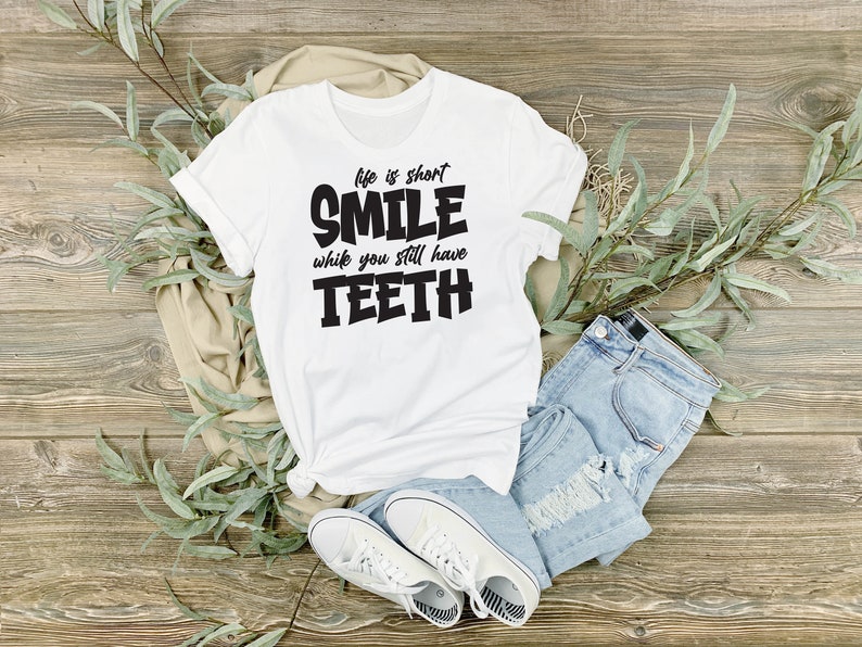 May include: White t-shirt with black text that reads "Life is short, Smile while you still have Teeth". The shirt is laid out on a wooden surface with a pair of jeans and white trainers.