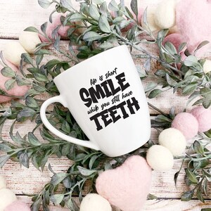 May include: White ceramic mug with a black graphic that reads "Life is short, Smile while you still have teeth". The mug is surrounded by pink and white pom-poms and green leafy branches.
