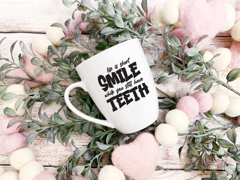 May include: White ceramic mug with a black graphic that reads "Life is short, Smile while you still have teeth". The mug is surrounded by pink and white pom-poms and green leafy branches.