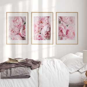 May include: Three framed prints of pink peonies, arranged in a row on a white wall above a bed with white bedding and a gray throw blanket.