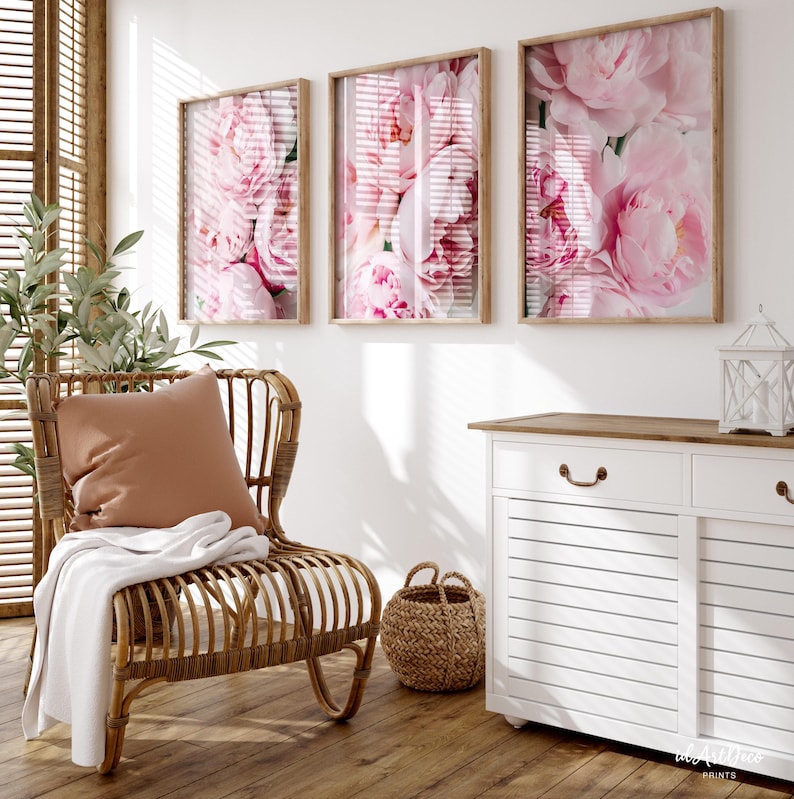 May include: Three framed floral prints with pink peonies on a white wall above a white dresser with a wooden top. A wicker chair with a brown cushion and a white throw blanket is in the foreground.