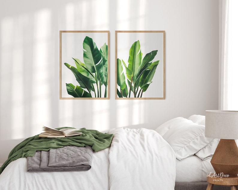 May include: Two framed prints of green tropical leaves, hanging on a white wall above a white bed with a green throw blanket.