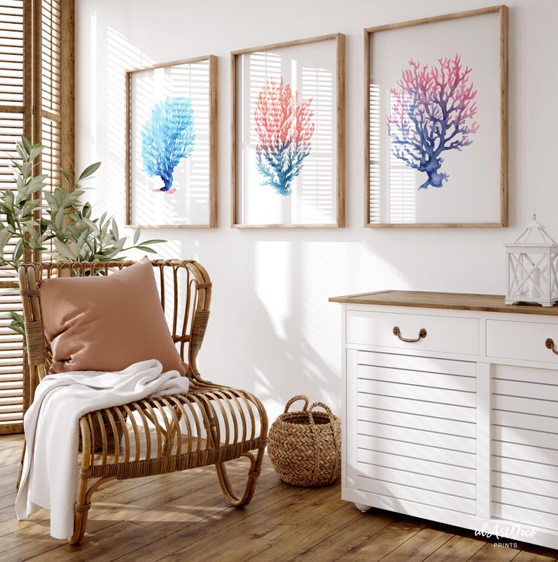 May include: Three framed watercolour paintings of coral, one blue, one pink, and one purple, hanging on a white wall above a white dresser with a brown top and a wicker chair with a brown cushion.
