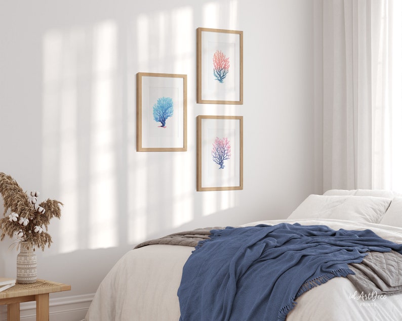 May include: Three framed watercolour prints of coral branches in shades of blue and pink, hanging on a white wall above a bed with a blue throw blanket.