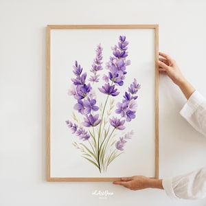 May include: A framed art print featuring a watercolor illustration of lavender flowers in shades of purple and green. The artwork is set against a white background and encased in a light brown wooden frame. The print is held by a person.