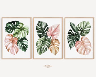 May include: A set of three framed prints showcasing watercolour illustrations of monstera leaves. The leaves are in shades of green, pink, and tan, set against a white backdrop. The frames are a light wood colour.